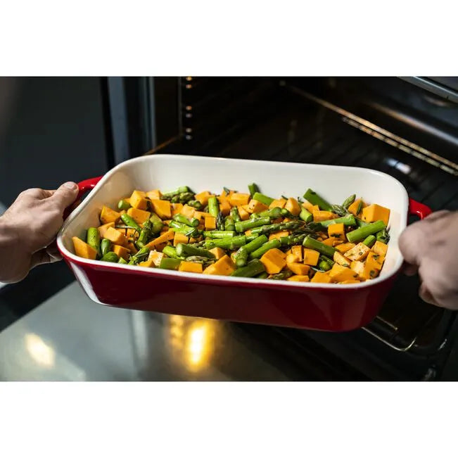 Person holding a red baking dish with vegetables in front of an open oven.