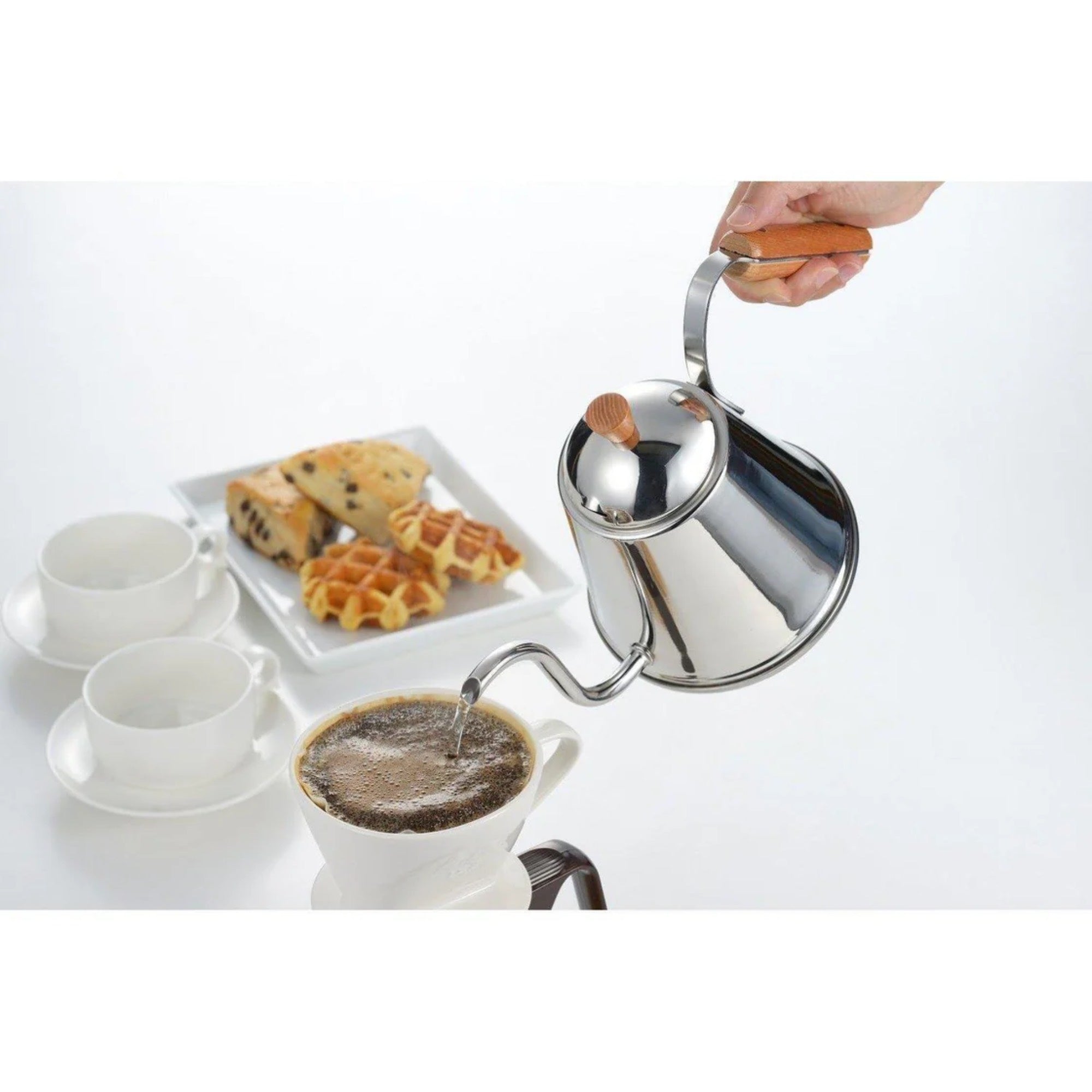 Stainless steel coffee pot being used to pour coffee into a white cup with waffles and a cookie on a white plate.