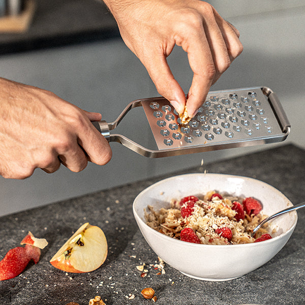 A stainless steel handheld microplane cheese grater being used to grate food over a white bowl of cereal with raspberries, with a person's hands visible in the action.