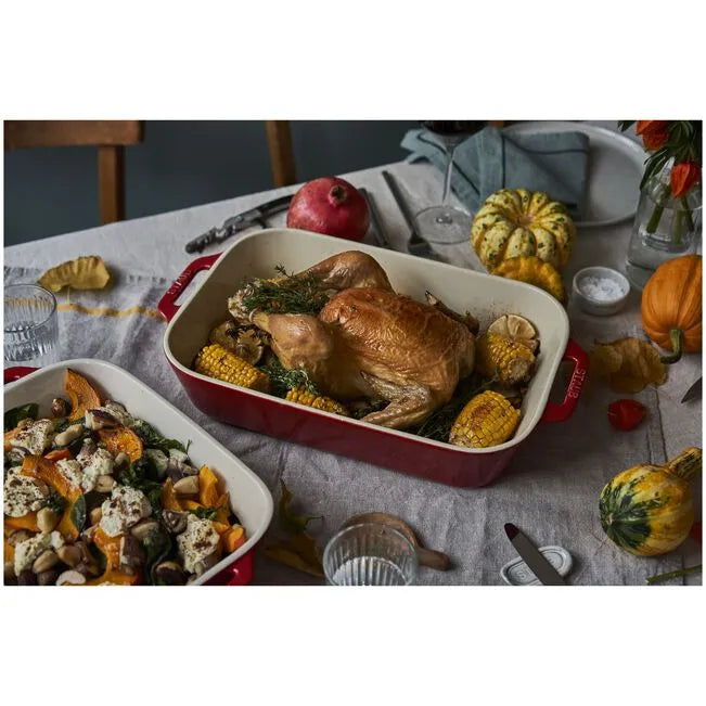 Roasted chicken with vegetables in a red baking dish on a table with pumpkins and a pomegranate.
