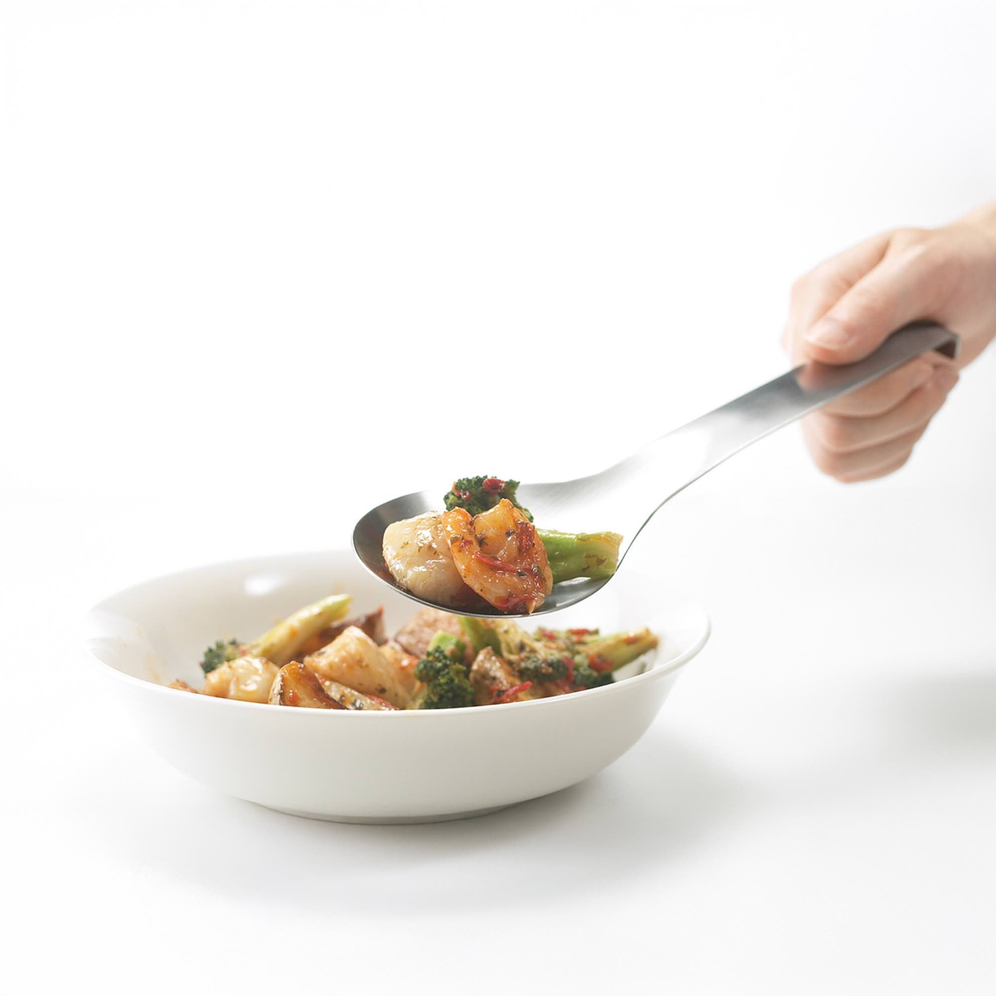 Eatoco stainless steel serving spoon lifting vegetables from a bowl, showcasing its sleek design and functionality.