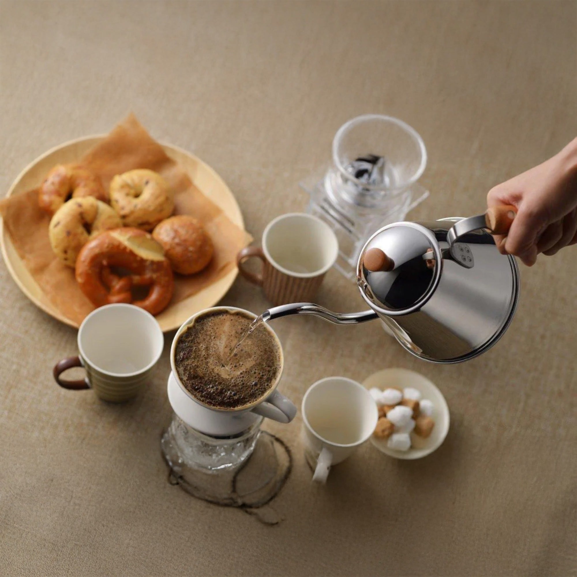 Person pouring coffee from a silver pour-over coffee maker into a white mug on a beige surface with pastries.