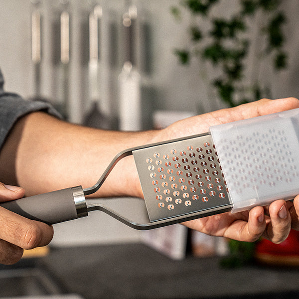 A close-up of a person holding a metal microplane grater with a gray handle and a translucent white plastic container, shown in a kitchen setting with blurred utensils and greenery in the background.